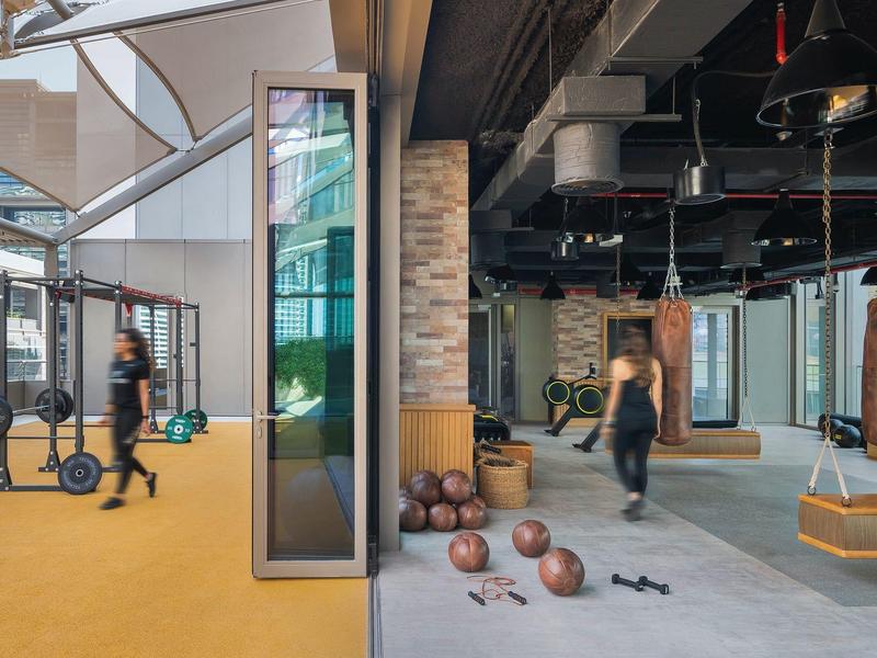 Moderner Fitnessraum mit orangenem Boden links und grauem Boden rechts, Sportgeräte und Menschen sichtbar.