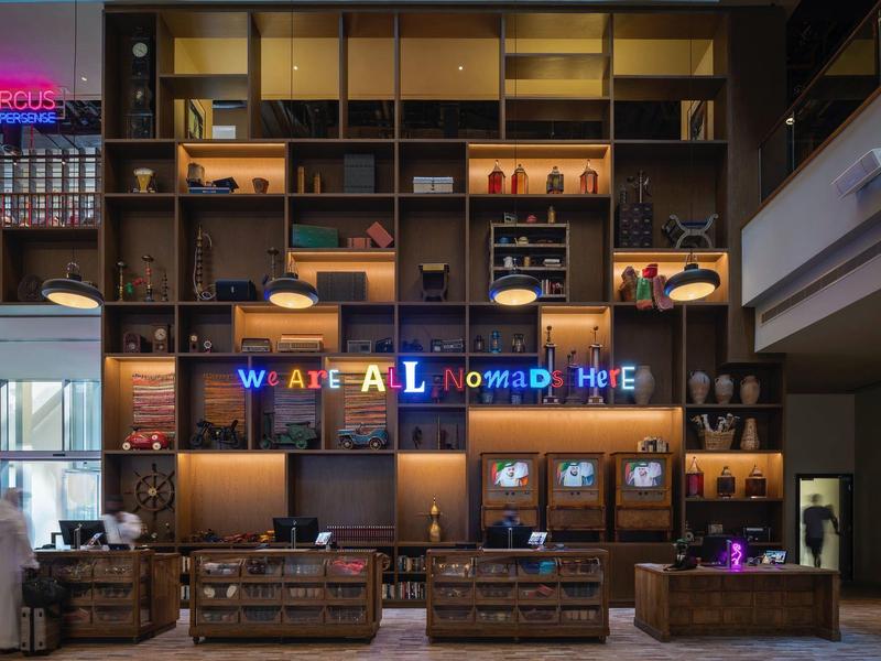 Moderne Hotellobby mit großen Holzregalen, Rezeption und blau-gelben Neonlichtern "WELCOME HOME".