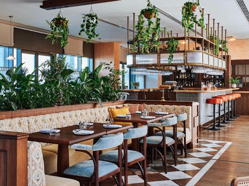 Helles Restaurant mit Holztischen, blauen Stühlen, Hängepflanzen und gemustertem Boden.