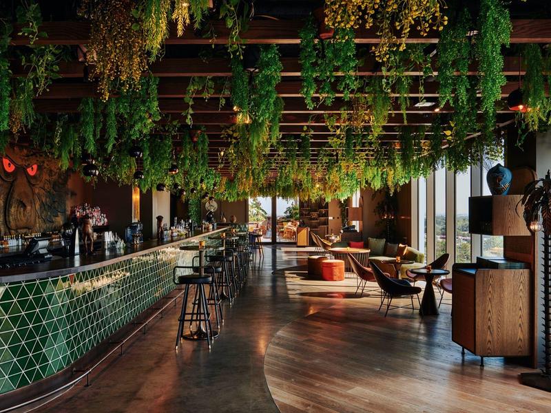 Modernes Café mit hängenden Pflanzen, grün gefliestem Tresen und gemütlichen Sitzbereichen.