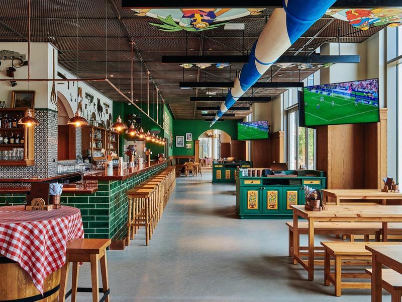 Moderner Gastraum mit Holz- und grünen Akzenten, großen Fenstern und bunt dekorierter Decke.