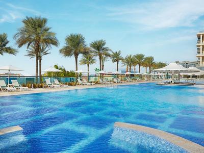 Großer Hotelpool mit Liegen, Palmen und Blick auf das Meer bei klarem Himmel.