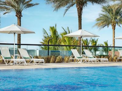 Poolbereich mit Liegestühlen, Sonnenschirmen und Palmen bei klarem Himmel.