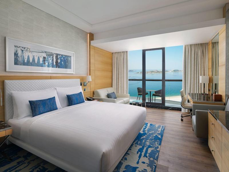 Modernes Hotelzimmer mit großem Bett, blauem Teppich, Schreibtisch und Meerblick durch Glasfenster.