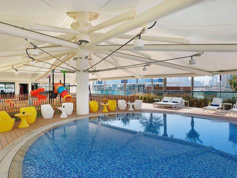 Runder Pool mit blauem Wasser unter großem Sonnenschirm und gelben Sesseln auf hellem Boden.