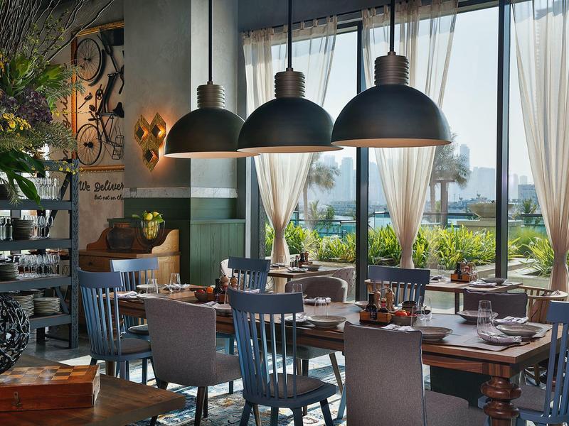 Gemütliches Restaurant mit Holztischen, blauen Stühlen, großen Fenstern und Blick auf grüne Pflanzen.