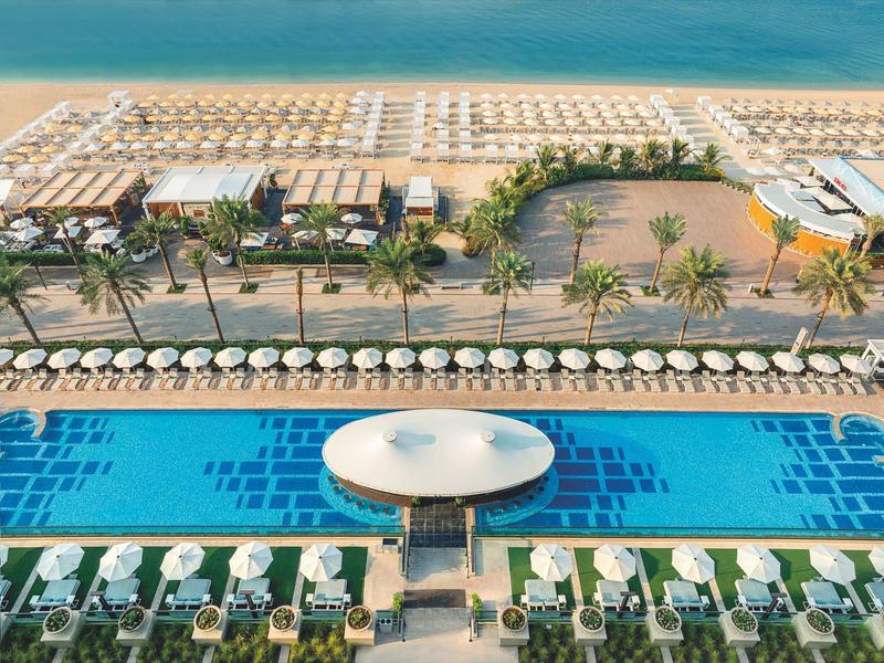 Blick auf ein großes Hotelpool mit Sonnenschirmen, Liegestühlen, Palmen und Strand im Hintergrund.