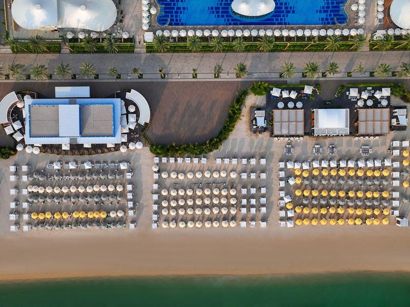 Luftaufnahme eines Strandes mit geordneten Sonnenschirmen, Liegen, Pools und Gebäuden.