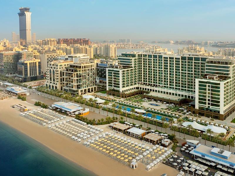Blick auf einen langen Sandstrand mit Reihen von Sonnenschirmen und großen modernen Hotels im Hintergrund.
