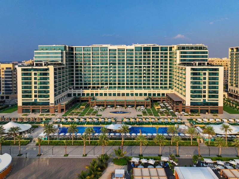 Großes modernes Hotel mit mehreren Stockwerken, Pools und Palmen davor bei klarem Himmel.