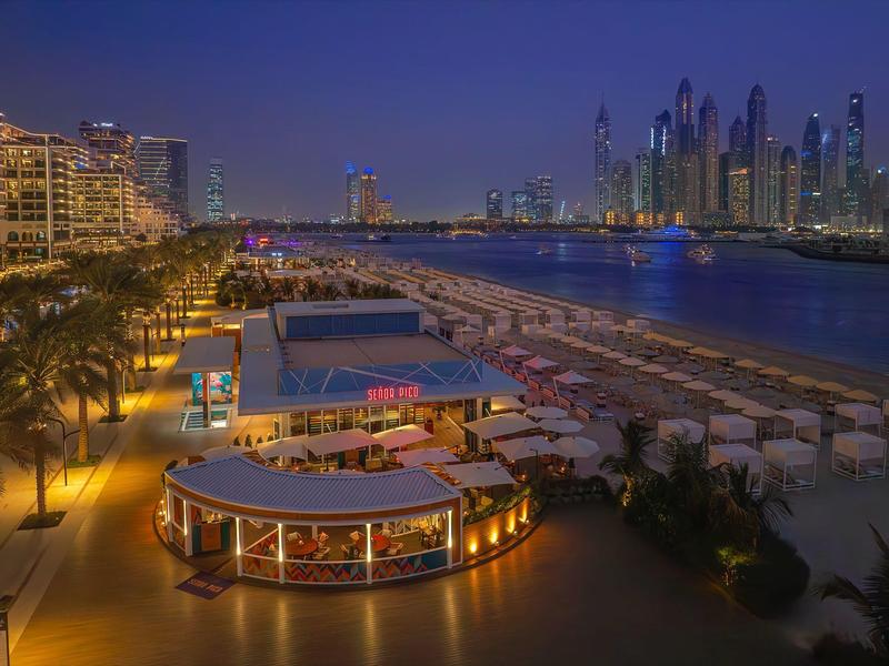Strandpromenade bei Nacht mit beleuchteten Gebäuden, Palmen und Strandliegen am Wasser.