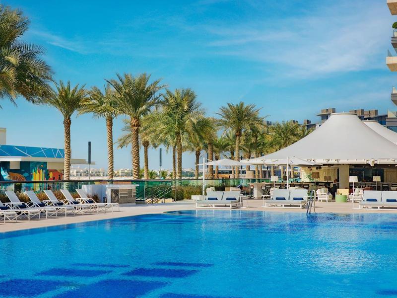 Großer Poolbereich mit Liegestühlen, Sonnenschirmen und Palmen unter klarem Himmel.