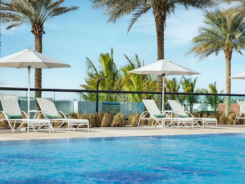 Poolbereich mit Liegestühlen, Sonnenschirmen und Palmen unter blauem Himmel.