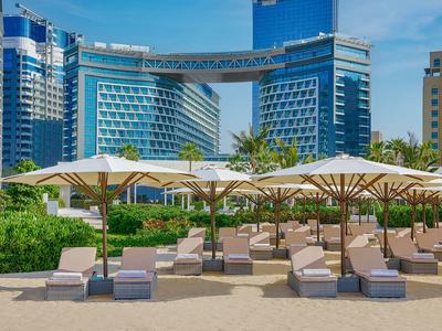 Strandliegen mit weißen Sonnenschirmen vor modernen, hohen blauen Glasgebäuden bei klarem Himmel.