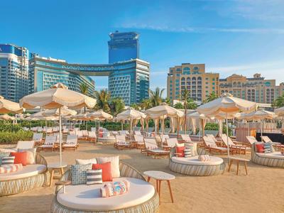 Strand mit weißen Liegen, Sonnenschirmen und hohen Gebäuden im Hintergrund bei klarem Himmel.