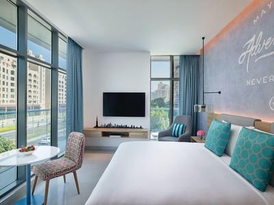 Nowoczesny pokój hotelowy z dużym łóżkiem, miejscem do siedzenia i oknami od podłogi do sufitu z widokiem na budynki.