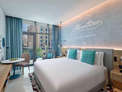 Helles Hotelzimmer mit großem Bett, blauen Vorhängen, runden Tischen und Fenster mit Stadtblick.