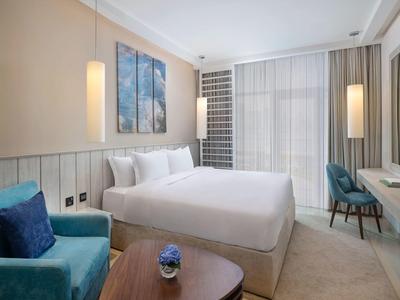 Modernes Hotelzimmer mit großem Doppelbett, zwei blauen Sesseln, Nachttischen und Fenster mit Vorhängen.