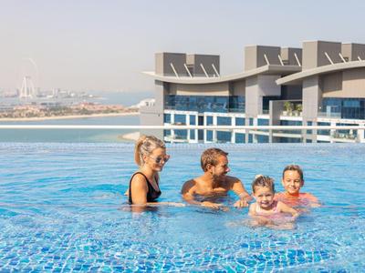 Vier Personen, darunter zwei Kinder, schwimmen und spielen in einem großen blauen Pool mit Stadtblick.