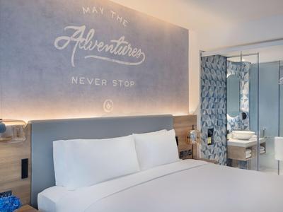 Nowoczesna przestrzeń wypoczynkowa hotelu z długimi stołami, tapicerowanymi ławkami i dekoracyjnymi półkami na ścianie.