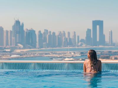 Frau im Pool blickt auf moderne Skyline mit hohen Gebäuden und blauem Himmel.