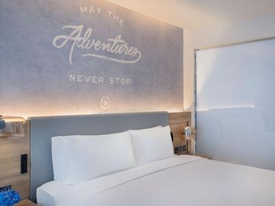 Nowoczesny pokój hotelowy z białym łóżkiem, drewnianą ścianą i motywującym cytatem nad zagłówkiem.