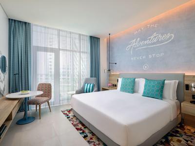 Nowoczesny pokój hotelowy z dużym łóżkiem, biurkiem i turkusowymi akcentami na poduszkach i zasłonach.