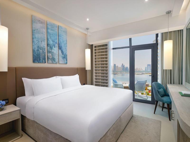 Modernes Hotelzimmer mit großem Bett, drei blauen Wandbildern und Fenster mit Stadt- und Wasserblick.