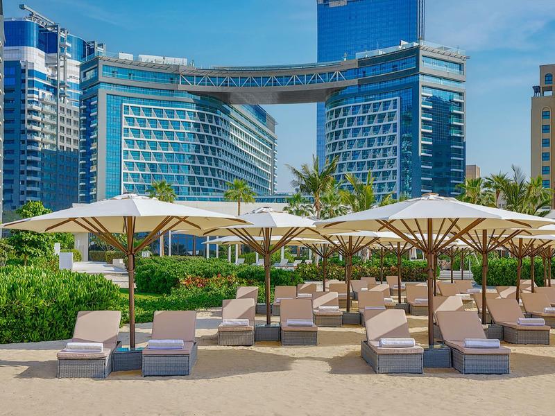 Strandliegen mit weißen Sonnenschirmen vor modernen, hohen blauen Glasgebäuden bei klarem Himmel.