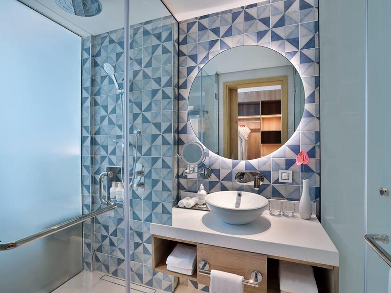 Modernes Badezimmer mit rundem Spiegel, weißem Waschbecken und geometrisch gemusterten Fliesen.