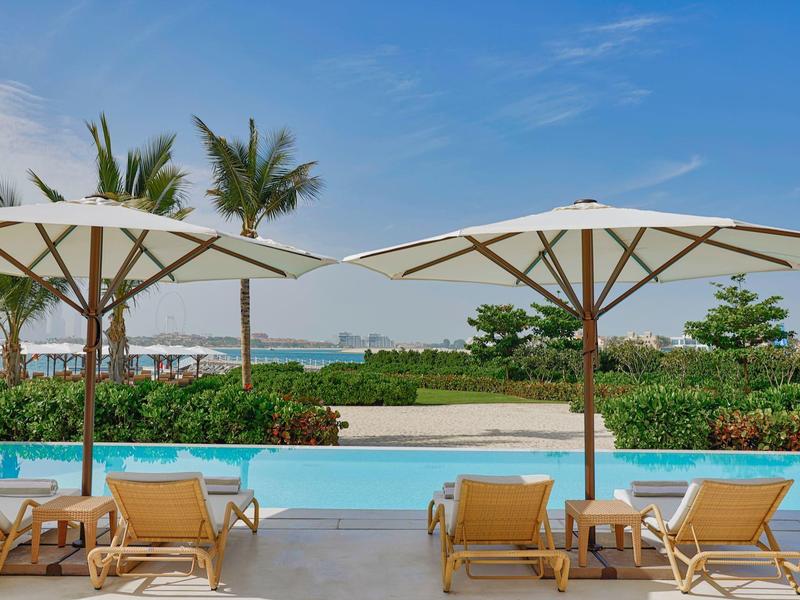 Poolbereich mit Liegestühlen und Sonnenschirmen, Palmen und Meer im Hintergrund unter blauem Himmel.