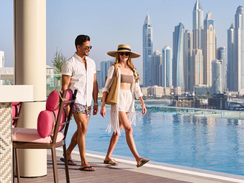 Ein Paar spaziert am Pool mit Blick auf moderne Wolkenkratzer in einer sonnigen Stadt.