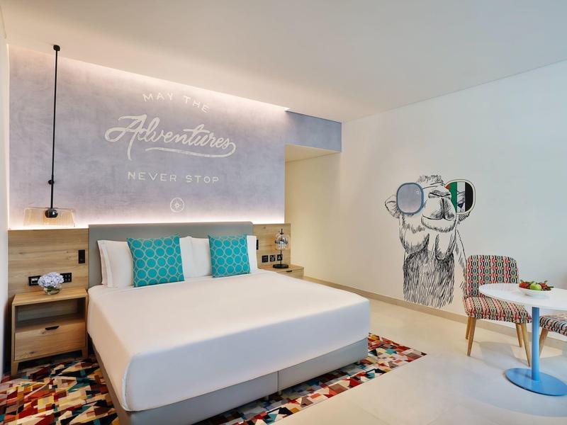 Modernes Hotelzimmer mit großem Bett, buntem Teppich, Wandkunst und Sitzecke mit Tisch und Stühlen.