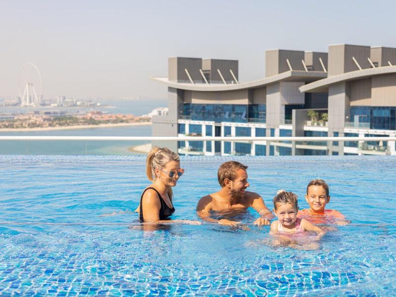 Vier Personen, darunter zwei Kinder, schwimmen und spielen in einem großen blauen Pool mit Stadtblick.