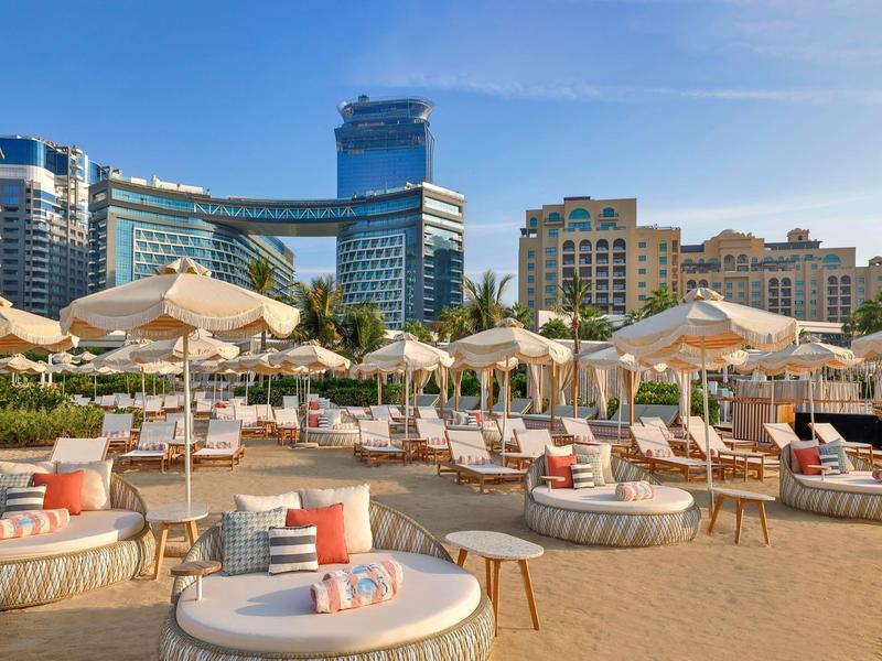 Strand mit weißen Liegen und Sonnenschirmen vor hohen modernen Gebäuden bei blauem Himmel.