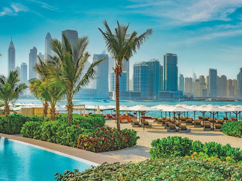 Städtische Poollandschaft mit Palmen, Sonnenschirmen und Hochhäusern im Hintergrund bei klarem Himmel.