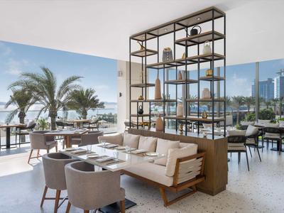 Nowoczesna część restauracyjna hotelu z sofą, krzesłami i widokiem na taras z palmami i miastem.