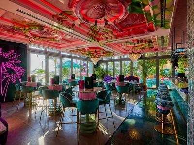 Kolorowy bar hotelowy z czerwonym sufitem, zielonymi krzesłami i dużymi oknami z widokiem na zewnątrz.