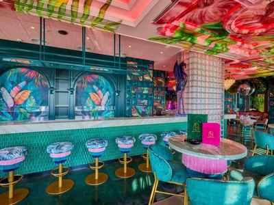 Kolorowy bar hotelowy z turkusowymi fotelami, stołkami barowymi i żywym kwiecistym sufitem.