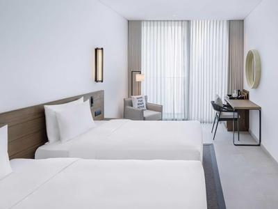 Helles, modernes Hotelzimmer mit zwei Einzelbetten, Schreibtisch, Stuhl und großer Fensterfront mit Vorhängen.