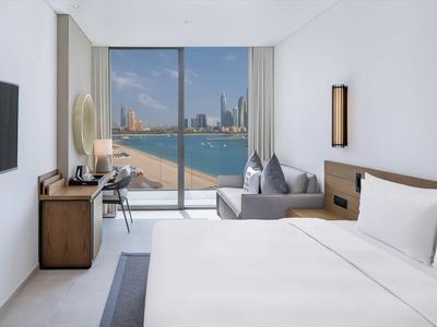 Hell eingerichtetes Hotelzimmer mit großem Bett, Sofa, Schreibtisch und Blick auf Wasser und Stadt.