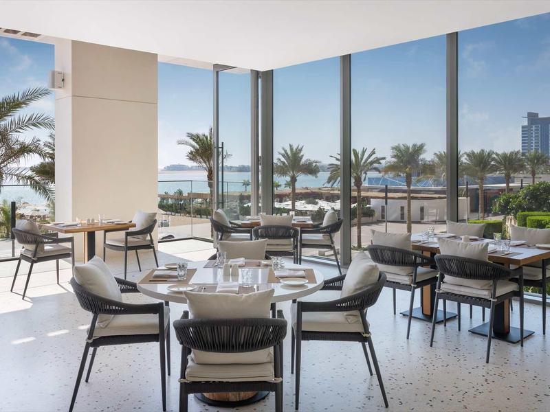 Moderner Speisesaal mit großen Fenstern, grauen Stühlen und Blick auf Palmen und Meer.