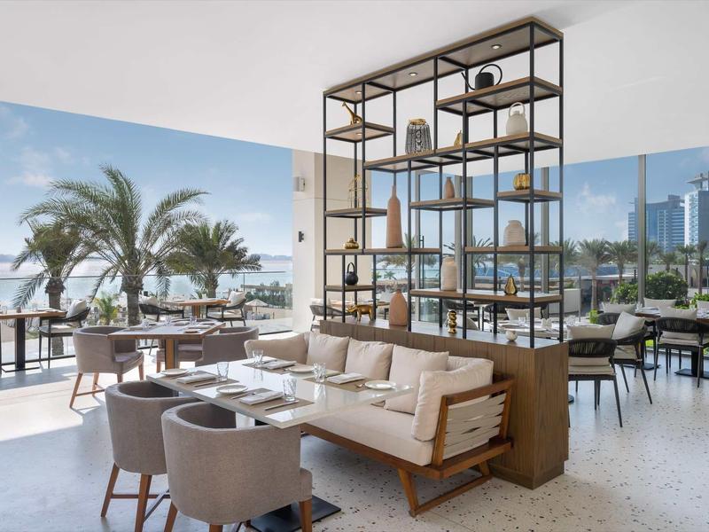 Helles Restaurant mit großen Fenstern, modernen Polstersesseln und regalförmiger Raumteiler.