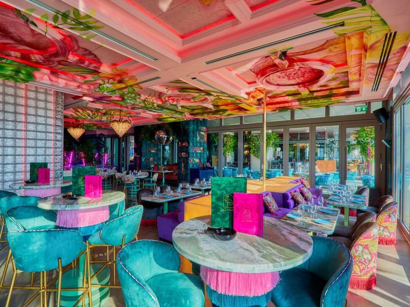 Buntes Restaurant mit türkisfarbenen Stühlen, rosa Tischen und floralen Deckendekorationen.