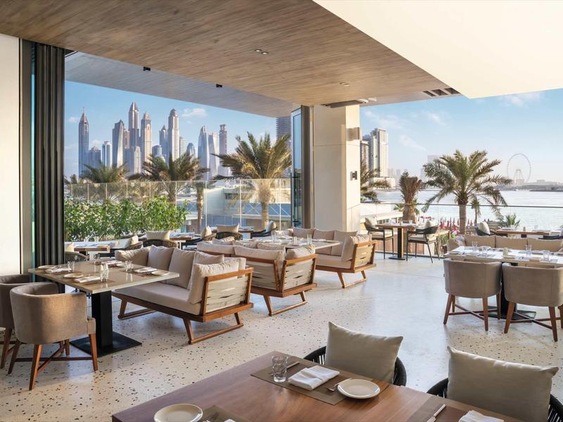 Freiluftrestaurant mit Holzmöbeln, weißen Tischen, Palmen und Blick auf eine moderne Skyline am Wasser.