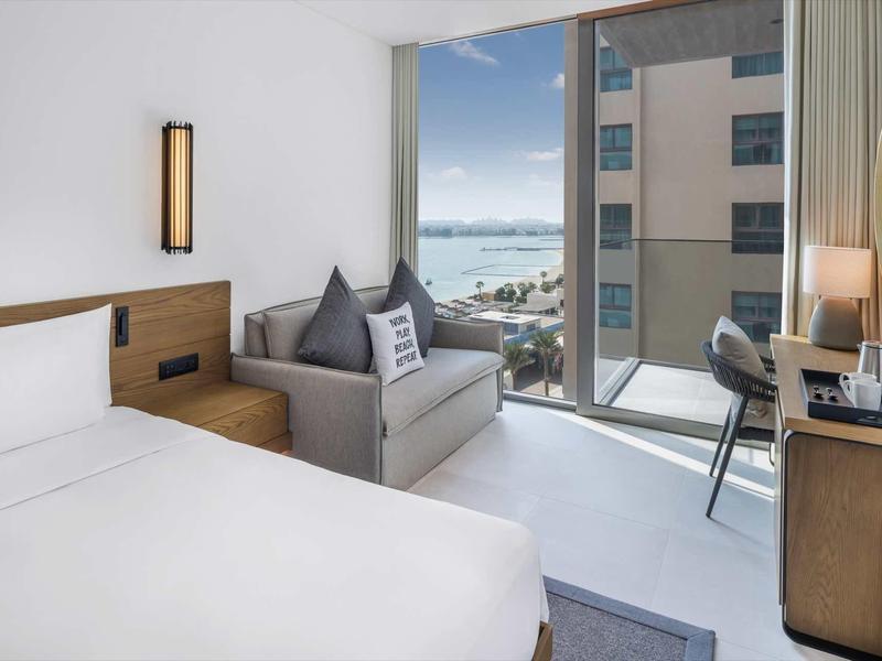 Helles Hotelzimmer mit Doppelbett, grauem Sofa, Schreibtisch und Meerblick durch große Fenster.