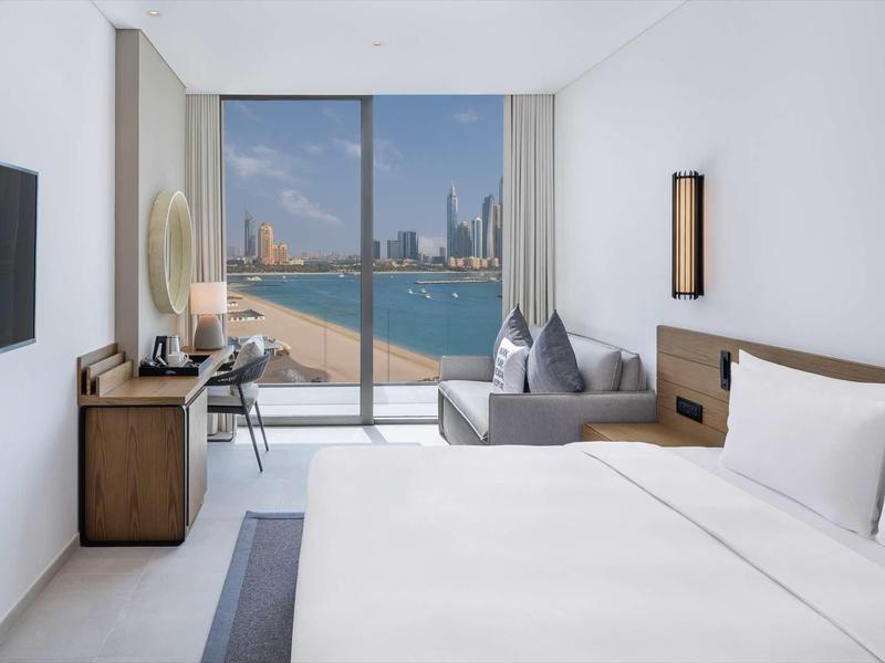 Hell eingerichtetes Hotelzimmer mit großem Bett, Sofa, Schreibtisch und Blick auf Wasser und Stadt.