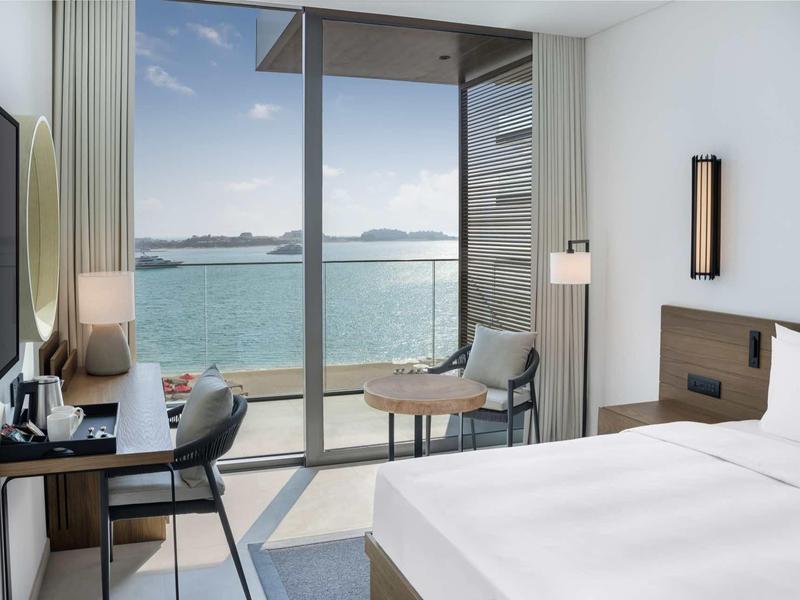 Modernes Hotelzimmer mit großem Bett, Schreibtisch und Meerblick durch bodentiefe Fenster.