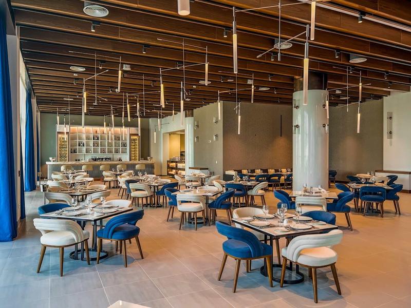 Großer, moderner Gastraum mit blauen Stühlen, Holztischen und Decke mit sichtbaren Holzbalken.
