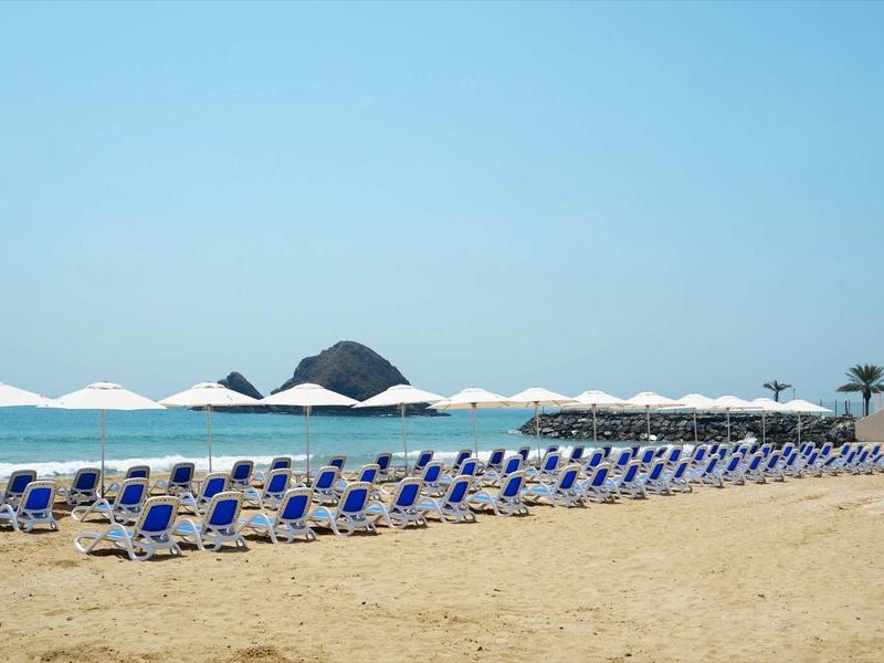 Reihe blauer Strandliegen mit weißen Sonnenschirmen auf hellem Sand vor blauem Meer und klarem Himmel.